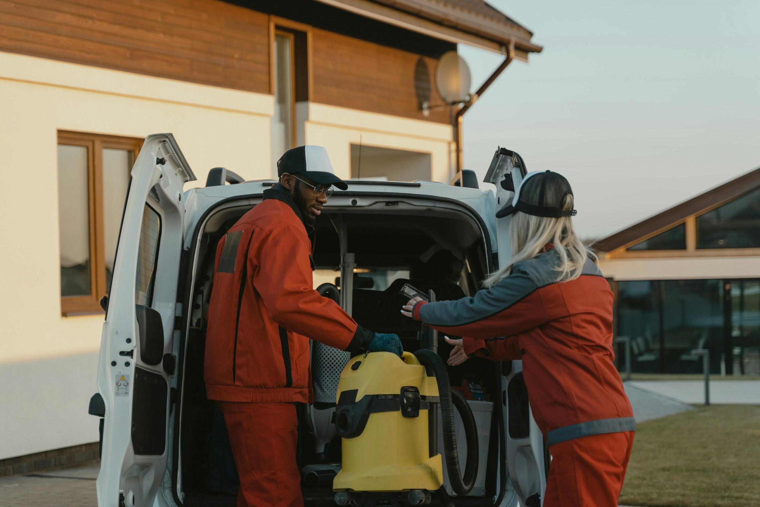 pexels-photo-6195895-6195895 Two cleaners in red uniforms load a vacuum into a white van outdoors.