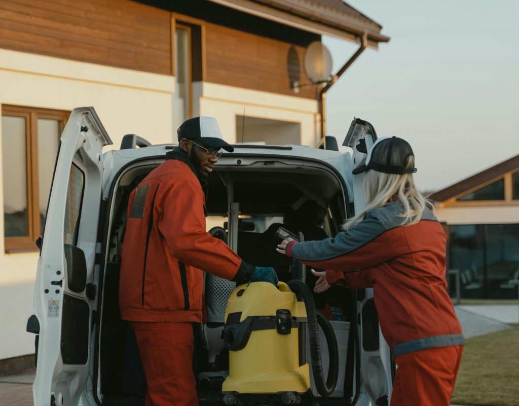 pexels-photo-6195895-6195895 Two cleaners in red uniforms load a vacuum into a white van outdoors.